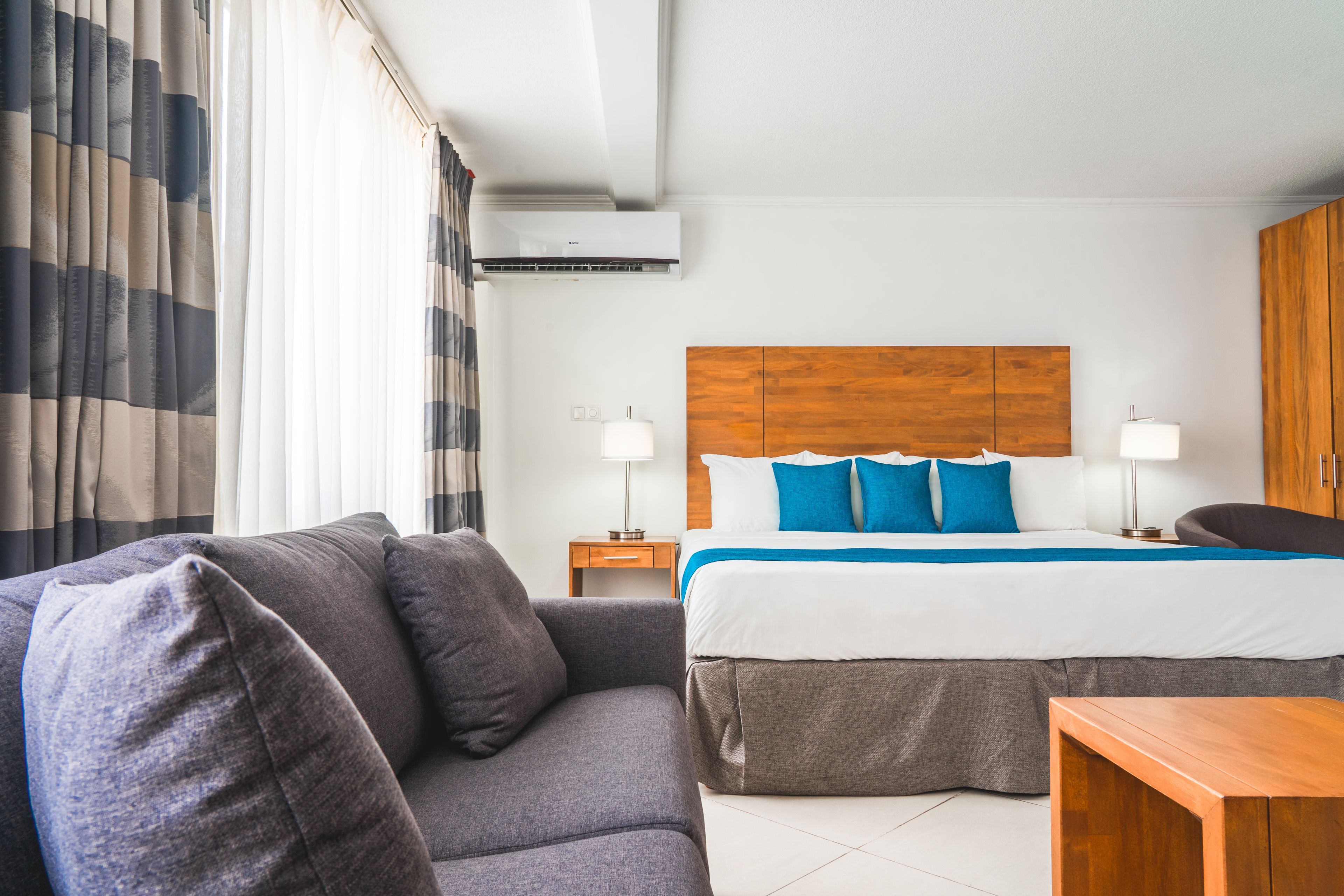 Una habitación de hotel moderna con una cama grande, un sofá gris, una mesita de noche de madera y un armario de madera.
