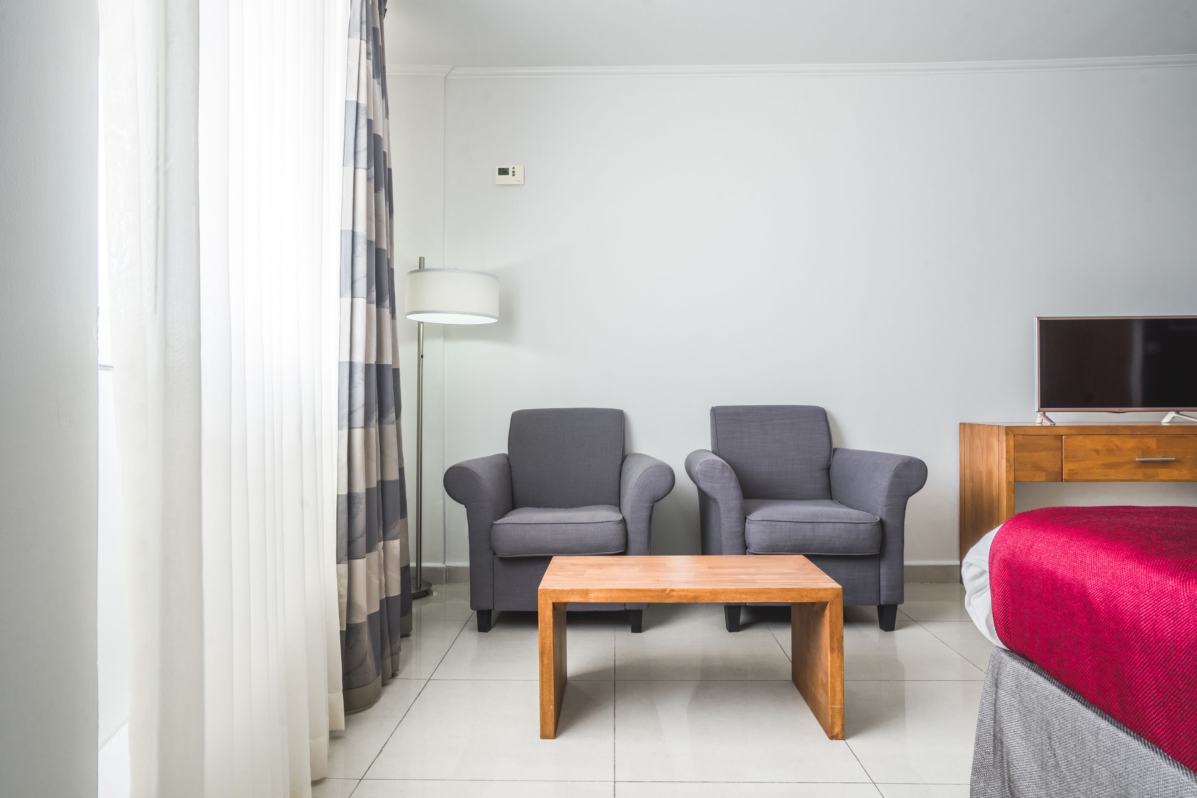 Een moderne hotelkamer met twee grijze fauteuils, een houten salontafel, een rode bed, een tv op een houten kast en een vloerlamp.