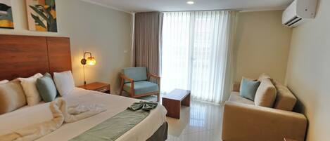 Habitación de hotel con cama, sillón, sofá, una mesita pequeña y un control remoto sobre una mesita de noche de madera.
