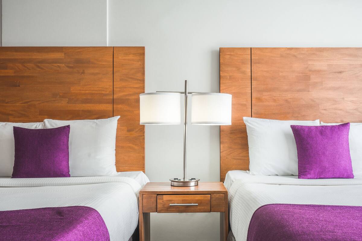 Habitación de hotel con dos camas, cada una con sábanas blancas y almohadas moradas, cabeceros de madera y mesitas de noche con lámparas.