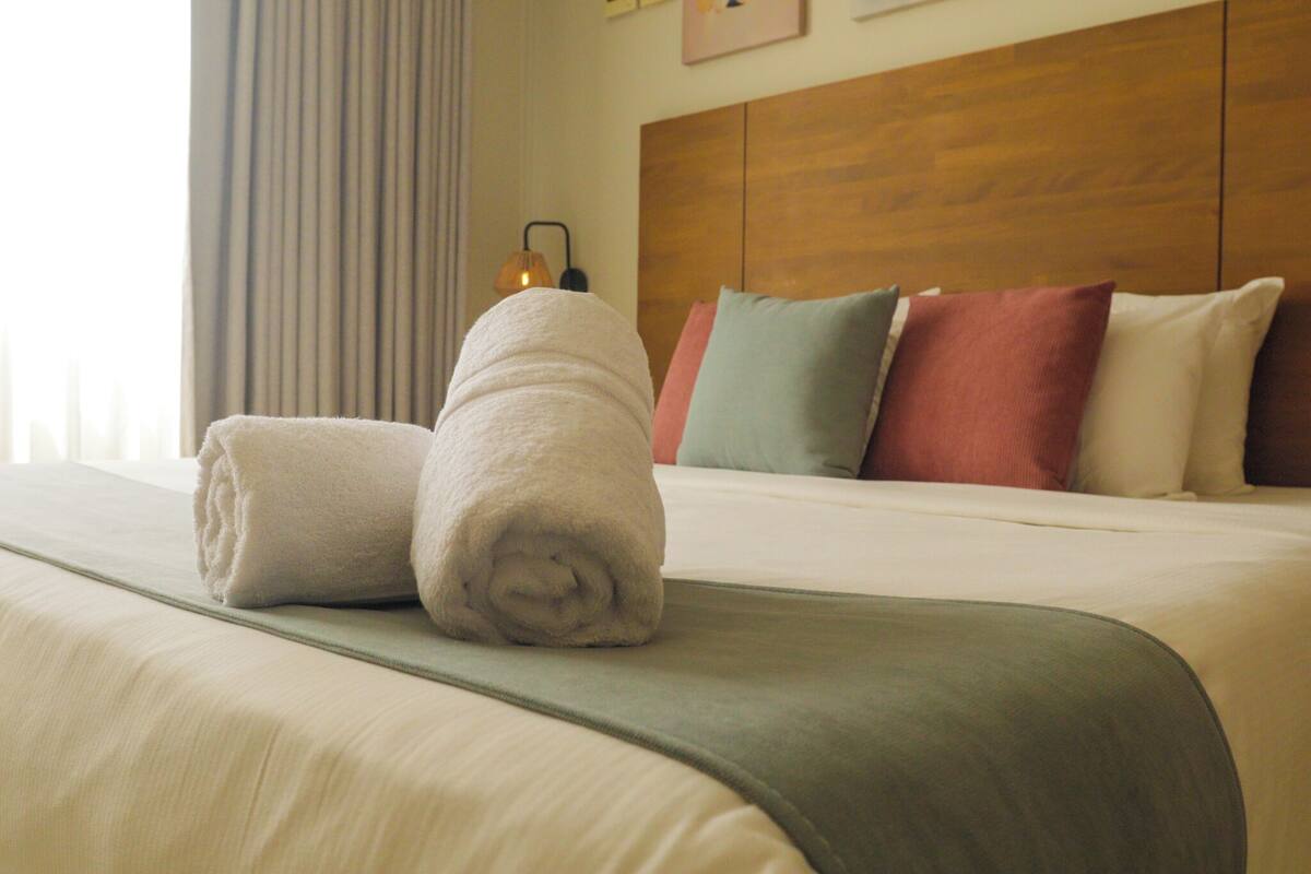 Una cama bien hecha con un edredón verde, cabecera de madera y varios cojines de distintos colores.