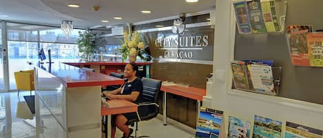 A hotel reception desk with a receptionist, a sign for city suites, and various travel brochures on the counter.
