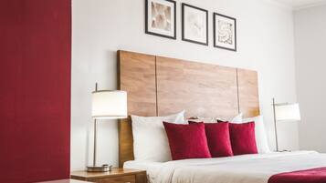 Een hotelkamer met een houten hoofdeinde, wit beddengoed, rode kussens en twee bedlampjes.