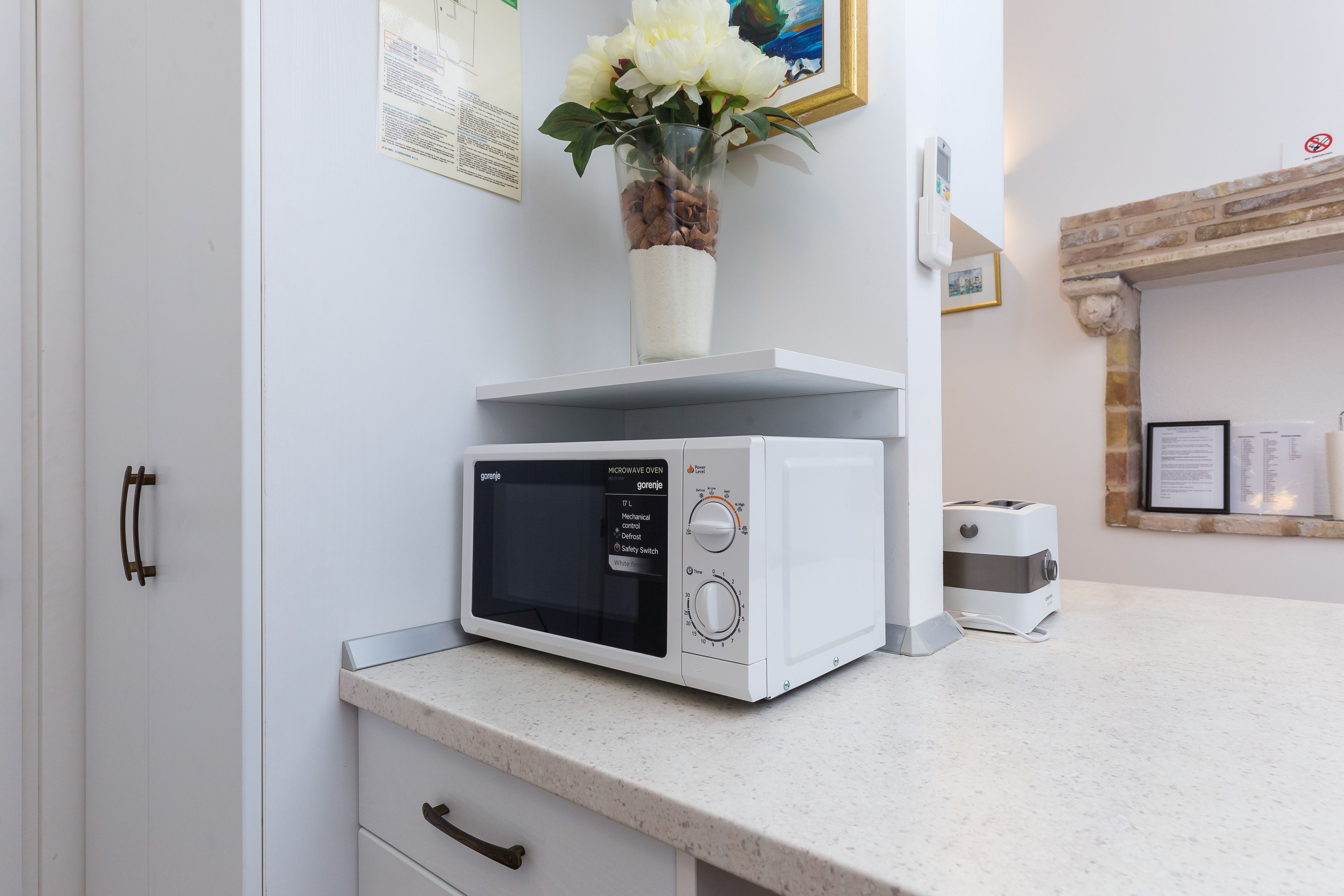 Eine Küche mit einer Mikrowelle auf der Arbeitsfläche, einem Toaster und einer Vase mit Blumen auf einem Regal.