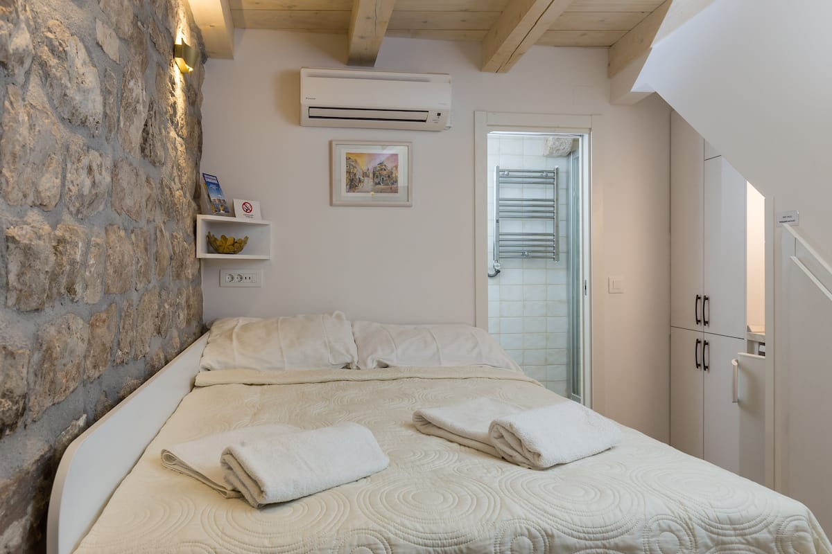 Une chambre avec un mur en pierre, un lit recouvert d’une courtepointe, un climatiseur fixé au mur, un tableau encadré et une porte dotée d’un porte-serviettes.