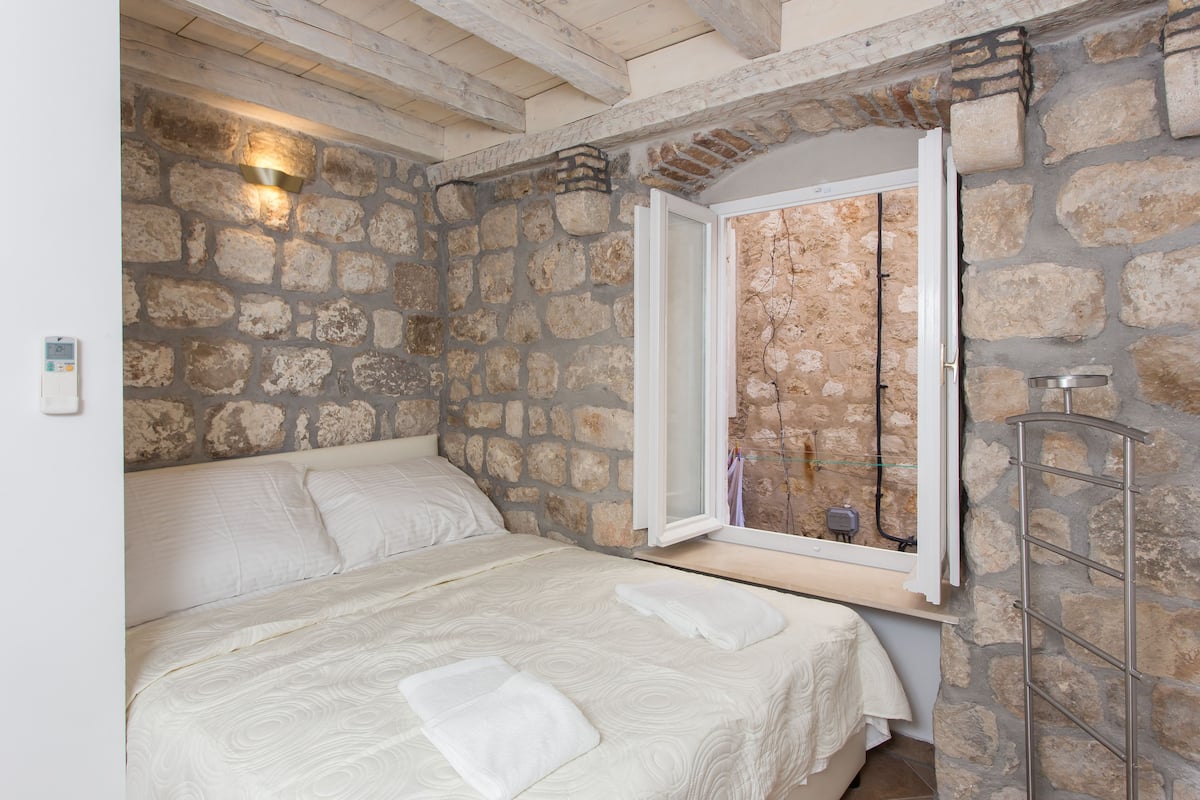 Une chambre avec un mur en pierre, un lit recouvert de draps blancs et une fenêtre donnant sur l’extérieur.
