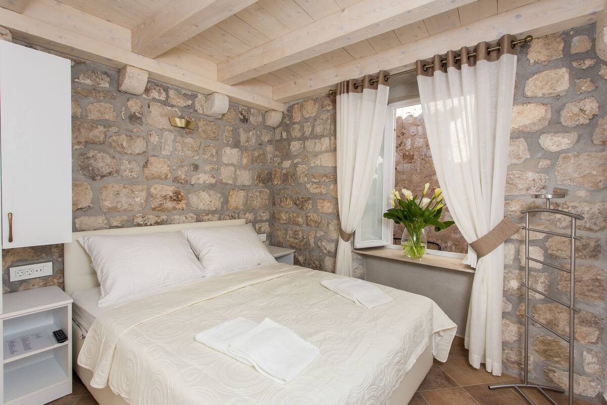Une chambre avec un mur en pierre, un lit recouvert de draps blancs, une table de chevet et une fenêtre avec des rideaux.