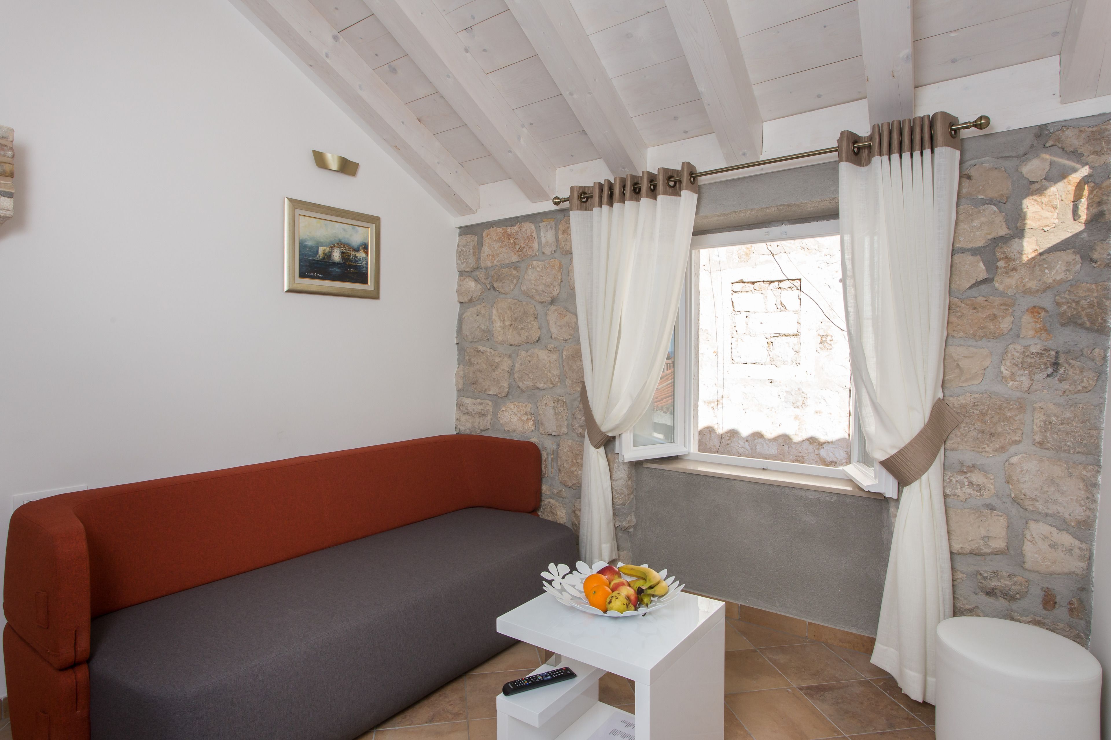 A room with a red sofa, a small white table with a fruit bowl, a remote control, and a stone wall.