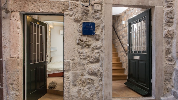 A stone building with two doors, one open leading to a room with a bed and a small dog, and another closed door with a staircase. - Apartments Bottega (Dubrovnik)