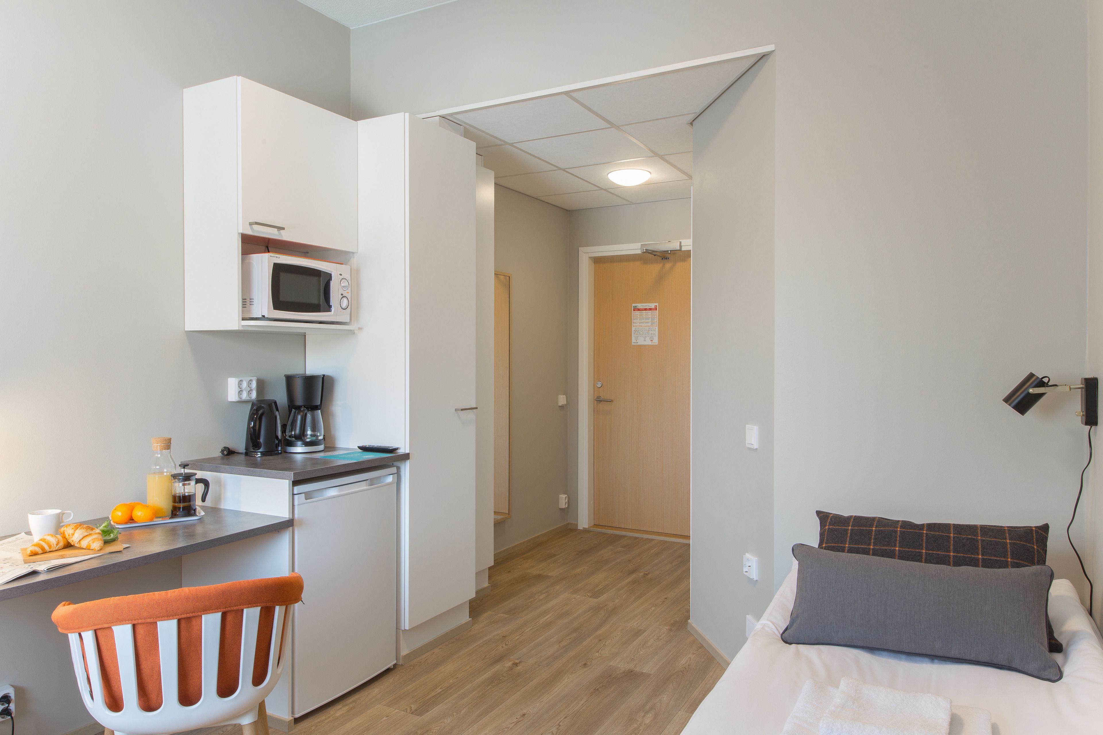 Une chambre d’hôtel compacte comprenant un coin cuisine, un lit avec des oreillers, une chaise et une petite table avec des articles pour le petit-déjeuner.