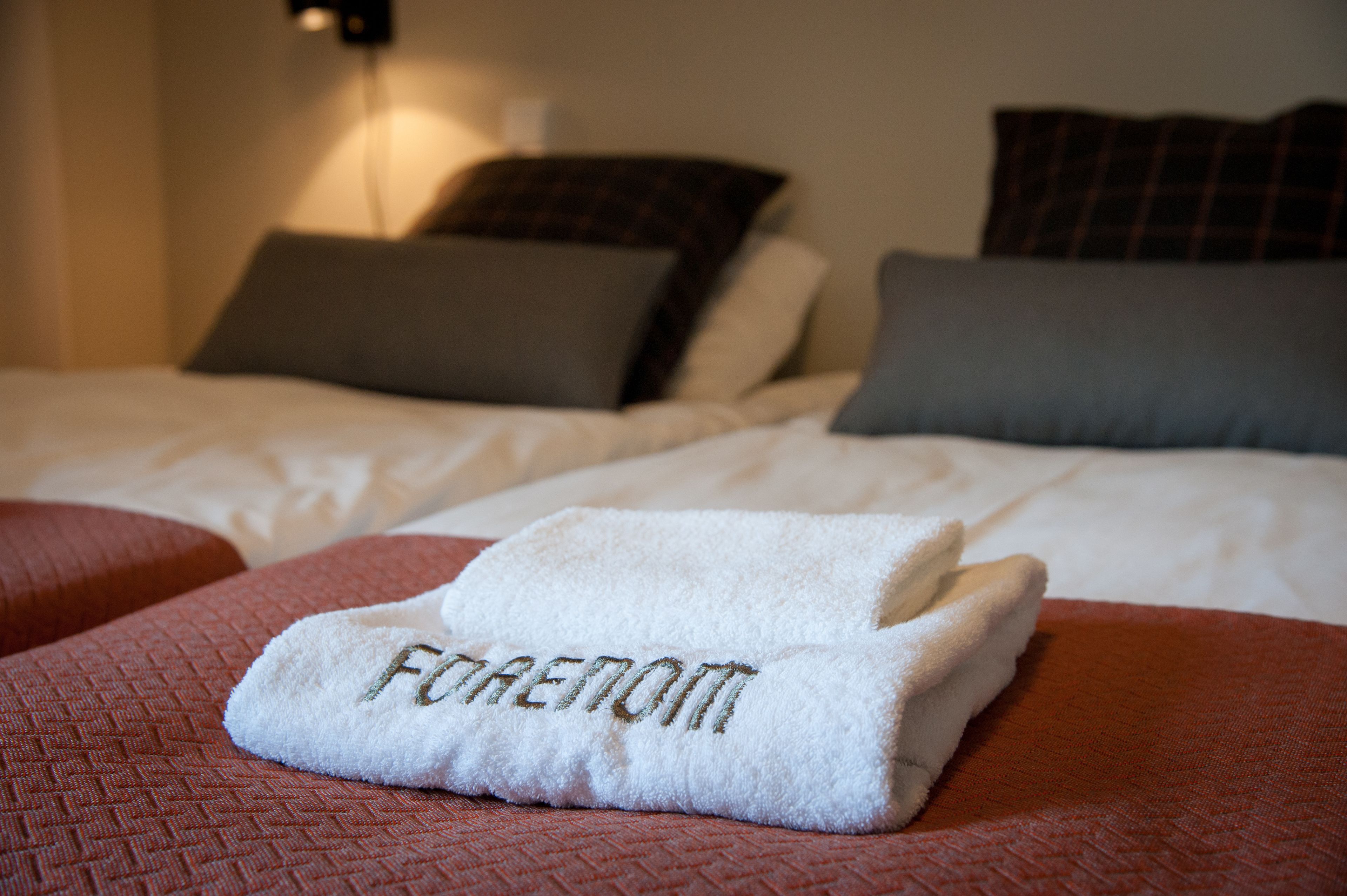 A neatly made bed with a folded white towel on a red bedspread, pillows in various colors, and a wall-mounted light fixture.