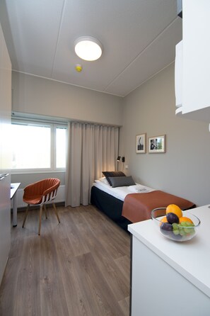 Habitación de hotel compacta con una cama individual, un escritorio, una silla y una mesita con fruta.