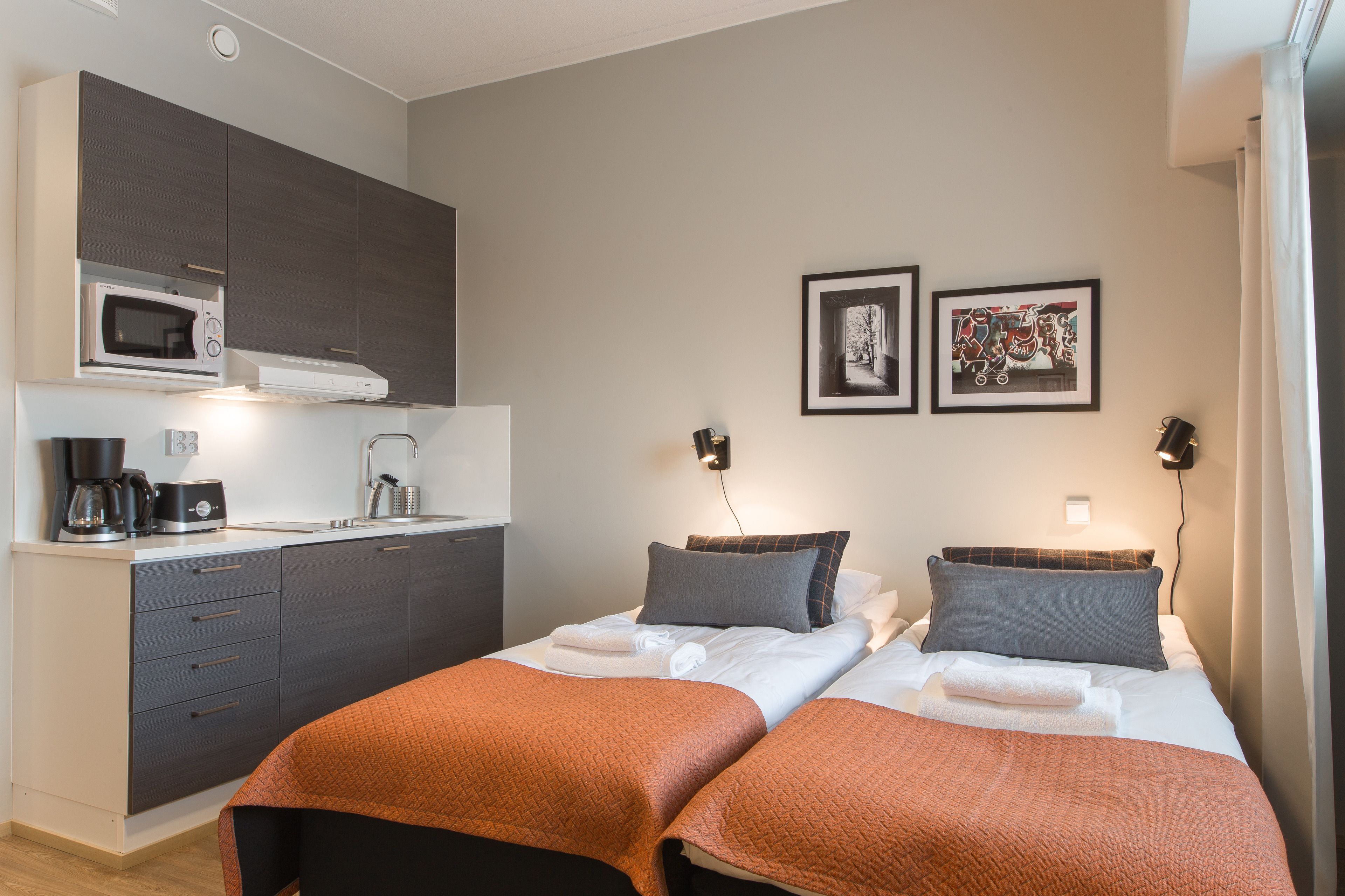 A modern hotel room with a kitchenette, two beds with orange bedding, and two framed pictures on the wall.