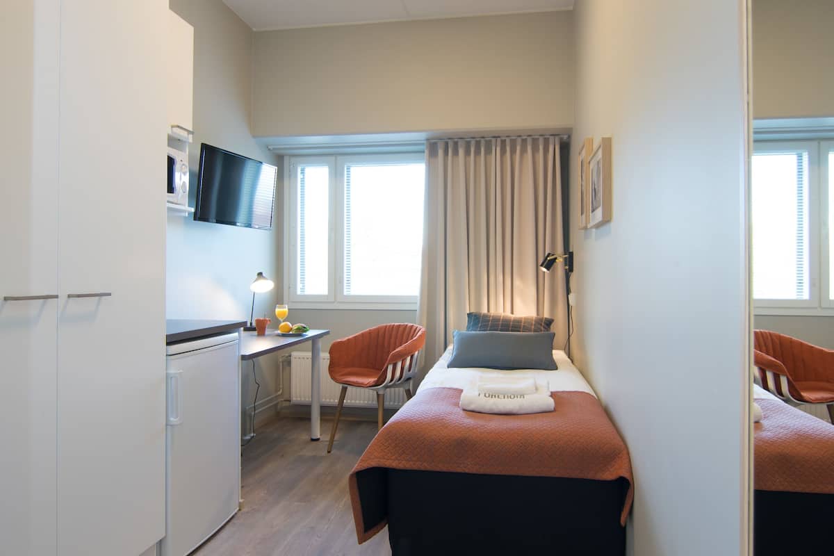 Ein kleines Hotelzimmer mit Bett, Schreibtisch, Stuhl, Fernseher und einem fensterbepflanzten Fenster.