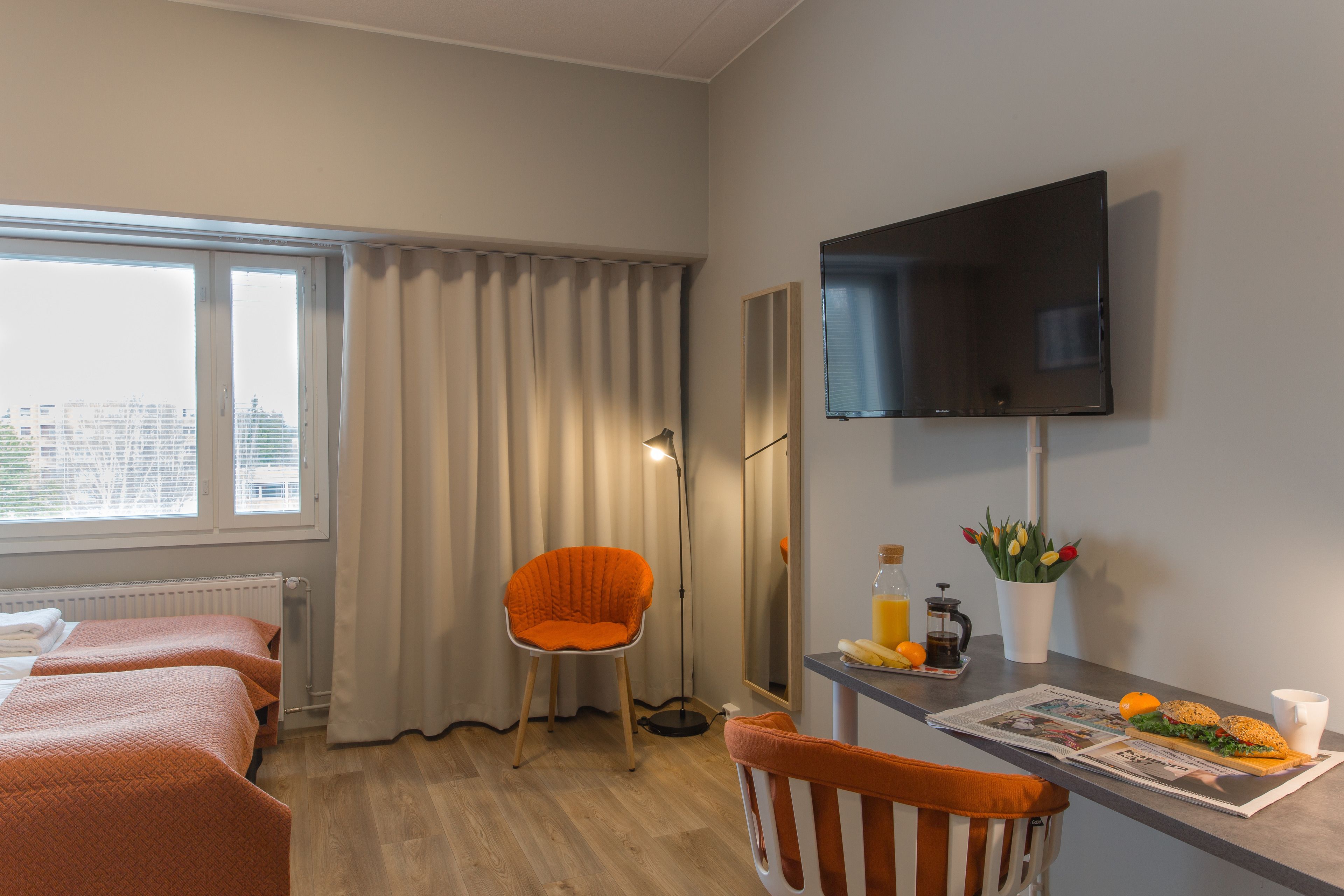 Ein Hotelzimmer mit einem Bett, einem Sessel, einem Schreibtisch mit Zeitung, einer Flasche und einer Vase mit Blumen.