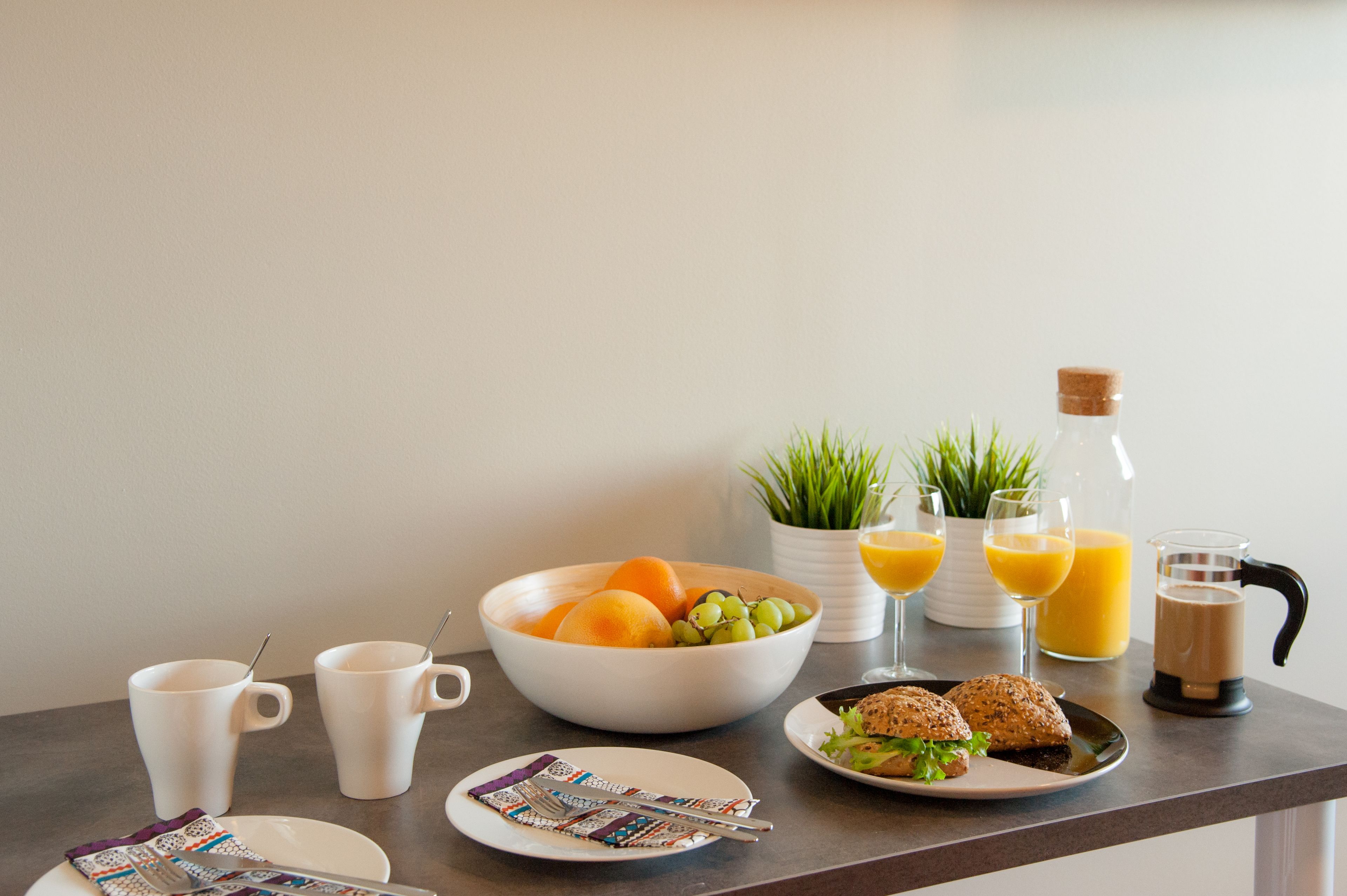 A breakfast table with a bowl of fruit, two cups, a bottle of orange juice, a glass of orange juice, a coffee maker, and a plate with a sandwich.