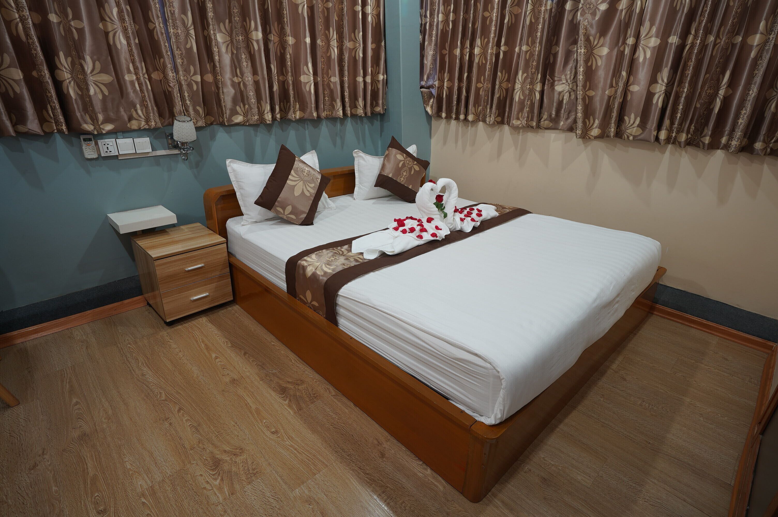 A neatly made bed with a white comforter and brown decorative pillows, a wooden nightstand with a phone and a small drawer, and a wall-mounted light fixture.