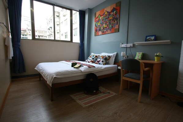 Habitación de hotel con cama, escritorio, silla, ventana con cortinas, un cuadro en la pared y un estante con objetos.