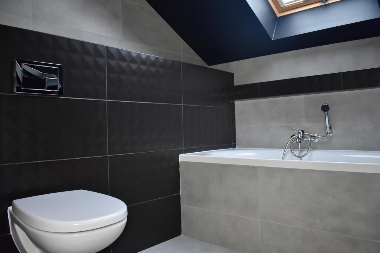 A modern bathroom with a white toilet, a bathtub with a showerhead, and dark tiled walls.