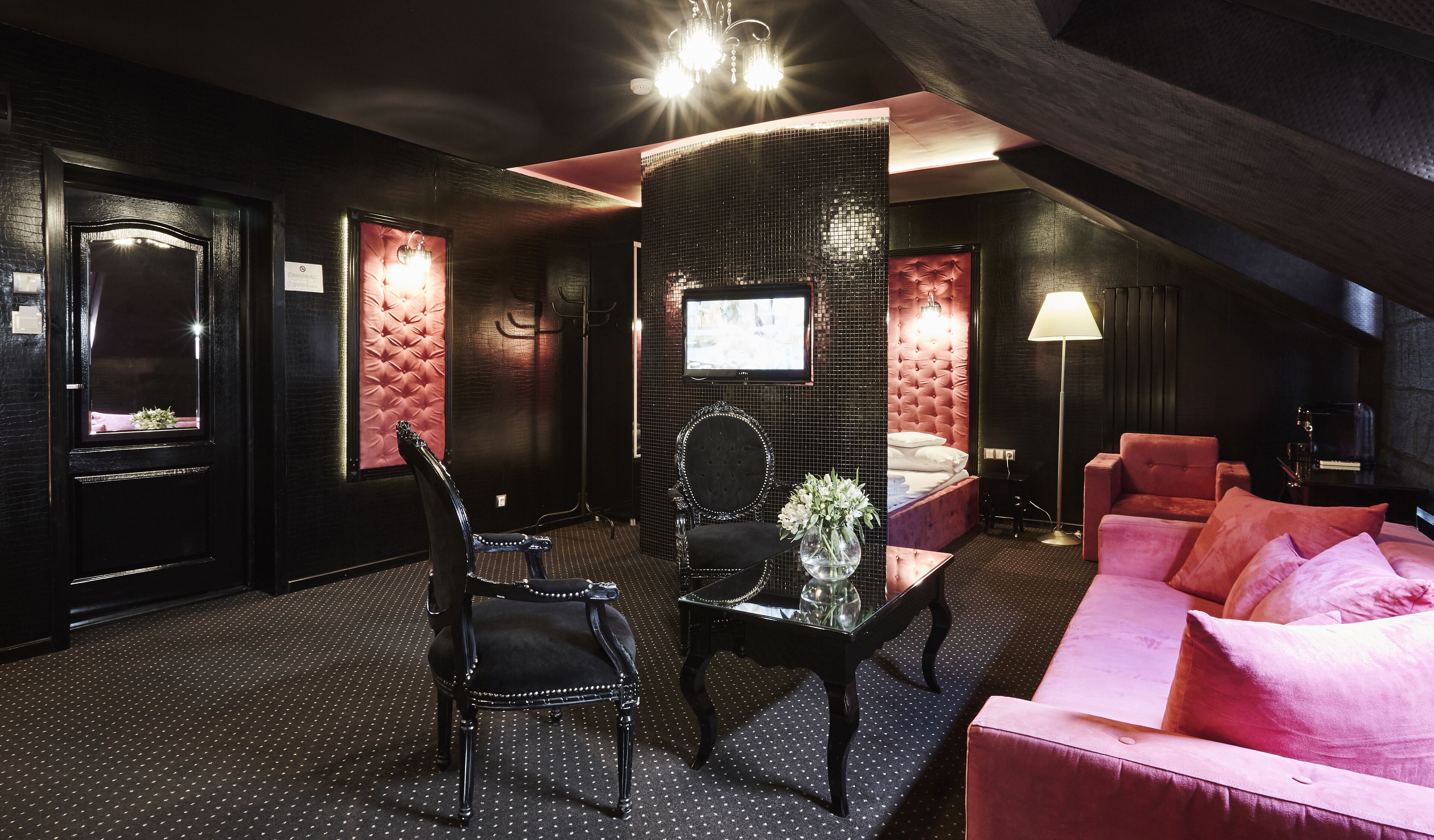 A modern hotel room with a dark color scheme, a tufted headboard, a large mirror, a television, a desk with a vase of flowers, and a pink sofa.