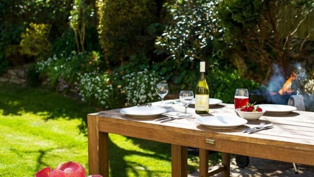 A wooden table set outdoors with plates, cutlery, a bottle of wine, and a glass of red drink. A pink cowboy hat is on the bench beside the table. - Gwithian Holidays, Godrevy House (Hayle)