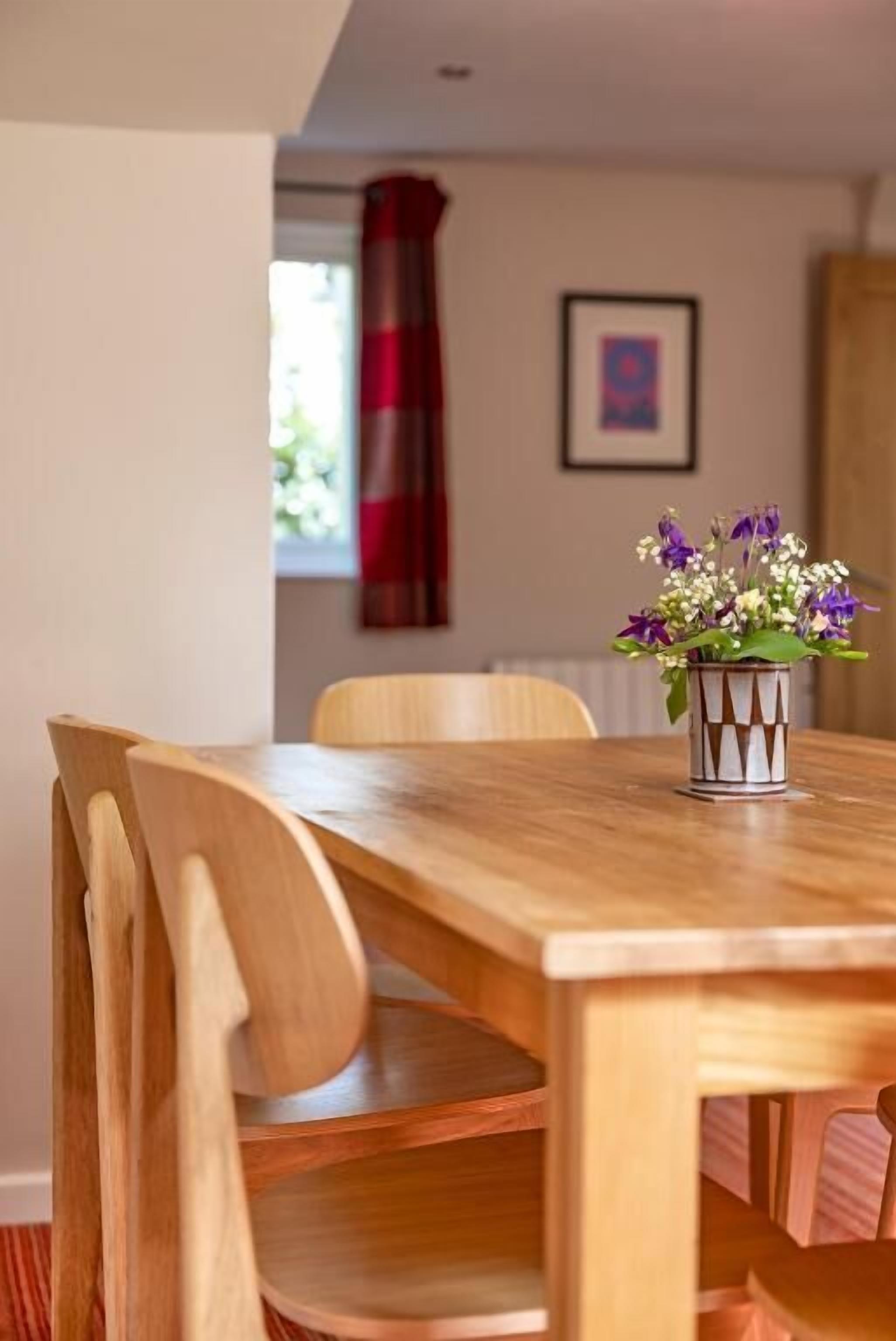 A wooden dining table with chairs, a vase with flowers, and a framed picture on the wall.