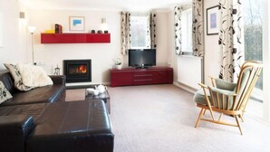 A living room with a fireplace, a television, a red entertainment unit, a sofa, a chair, and a window with curtains. - Gwithian Holidays, Godrevy House (Hayle)