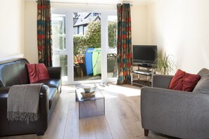 A living room with two sofas, a coffee table, a television, and a sliding glass door leading to a garden. - Gwithian Holidays, Godrevy House (Hayle)