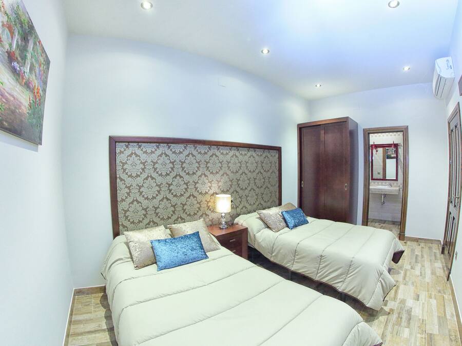 A hotel room with two beds, a nightstand with a lamp, a wooden wardrobe, and a bathroom visible through an open door.