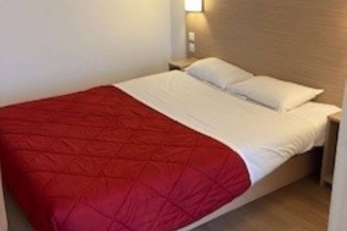 A single bed with a red blanket and white pillows in a room with wooden flooring and a nightstand.