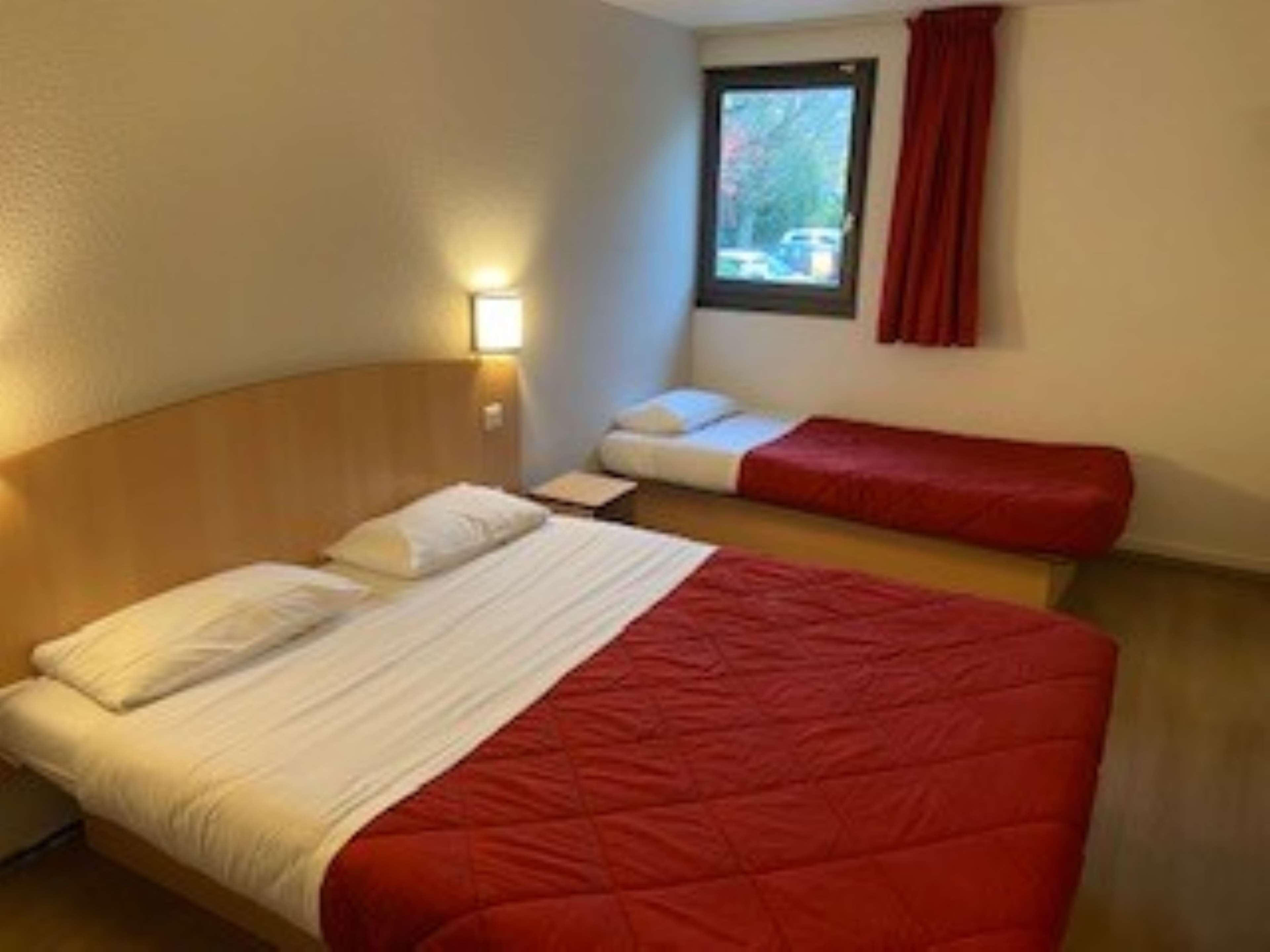 A hotel room with a large bed, a smaller bed, a window with curtains, and a wall-mounted light.