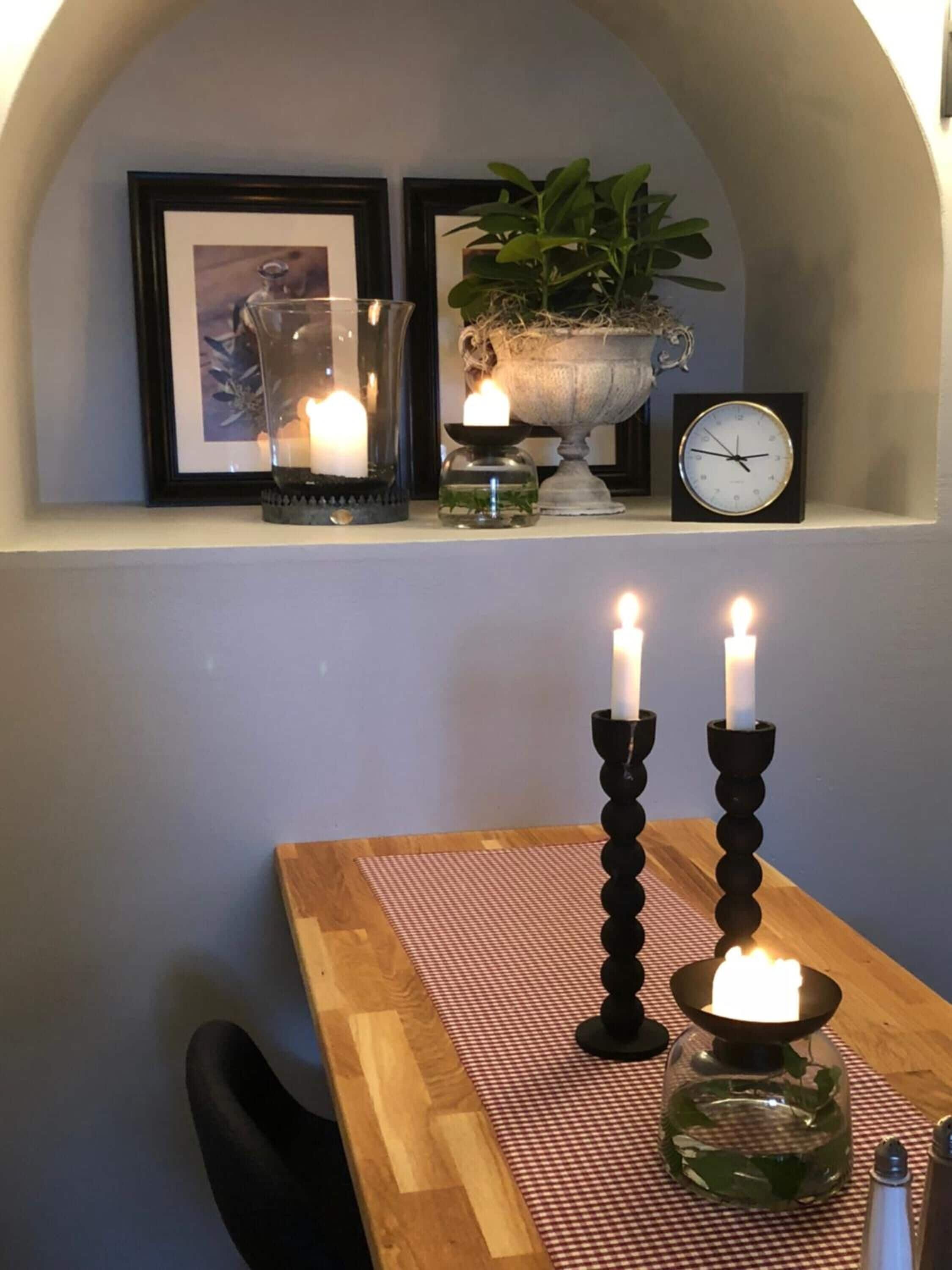 A cozy corner with a wooden table, two lit candles, a small vase with greenery, a framed picture, a potted plant, and a wall-mounted clock.