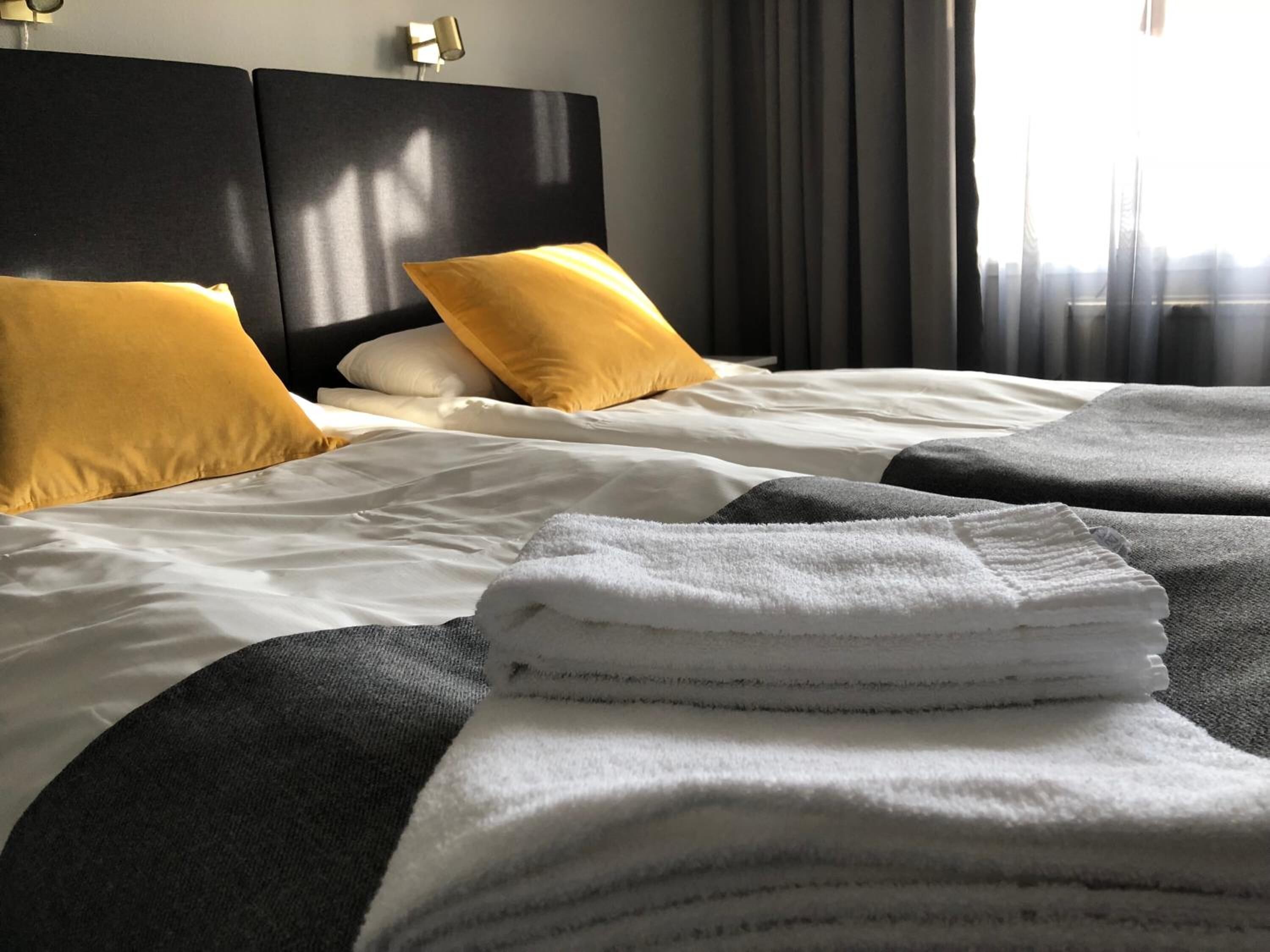 Una cama bien hecha con ropa de cama blanca y gris, dos almohadas amarillas y una pila de toallas blancas.