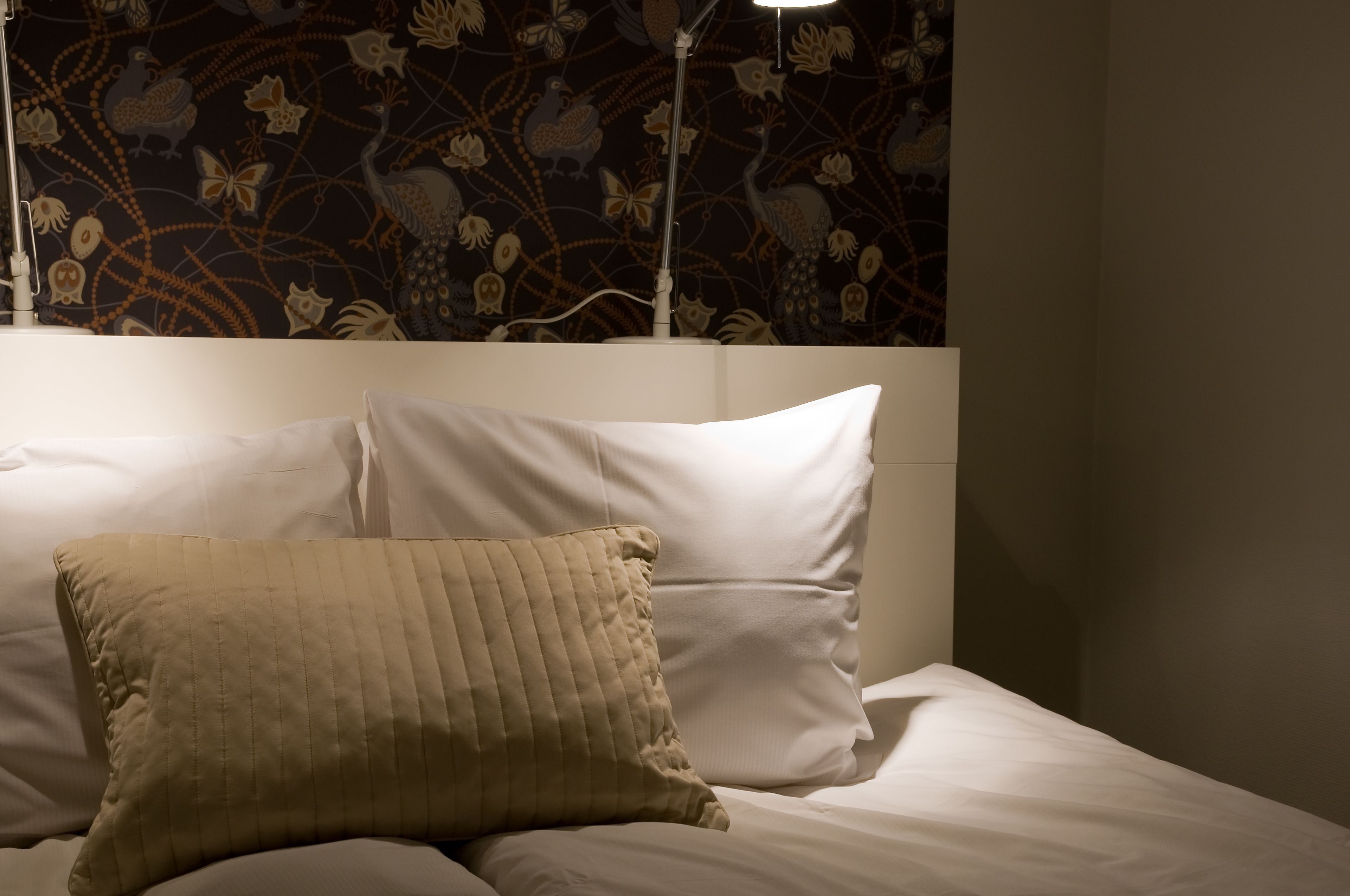 A bed with white linens and a beige pillow, a wall with a floral pattern, and a floor lamp.