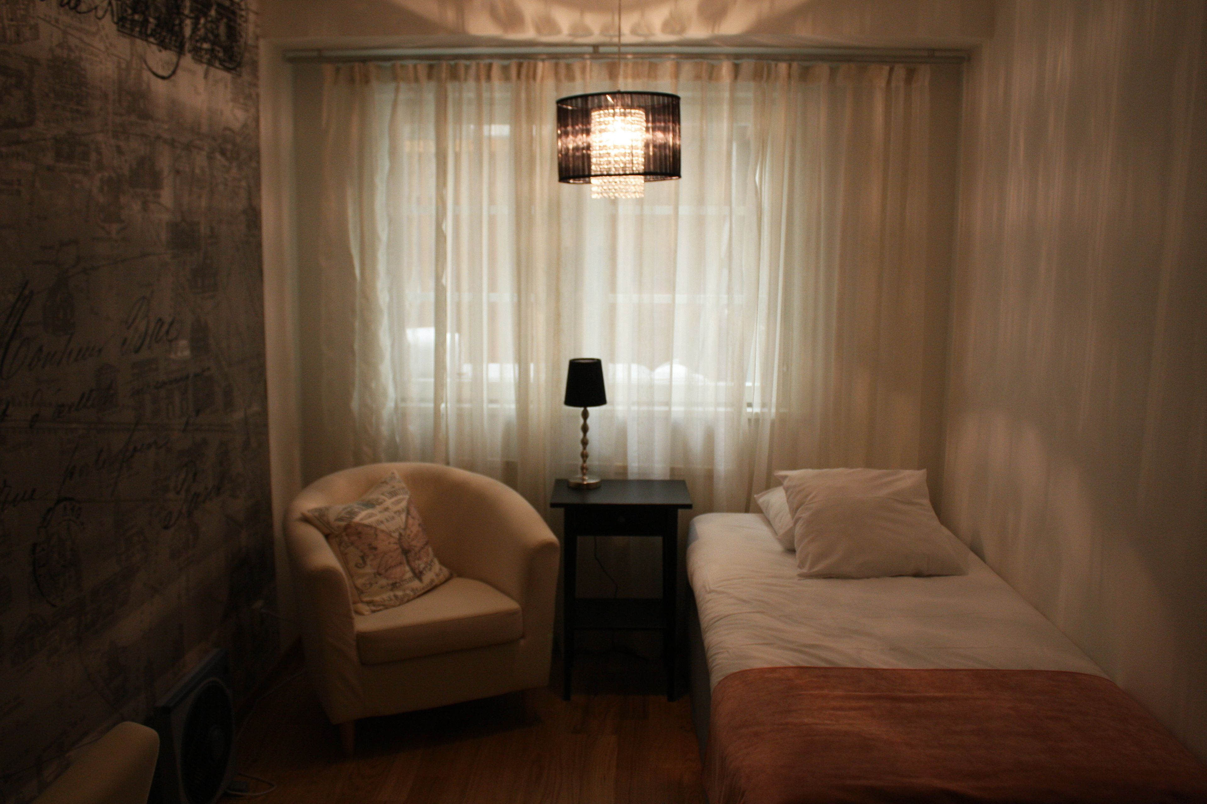 Ett hotellrum med en säng, en stol, ett litet bord med en lampa och tunna, genomskinliga gardiner.