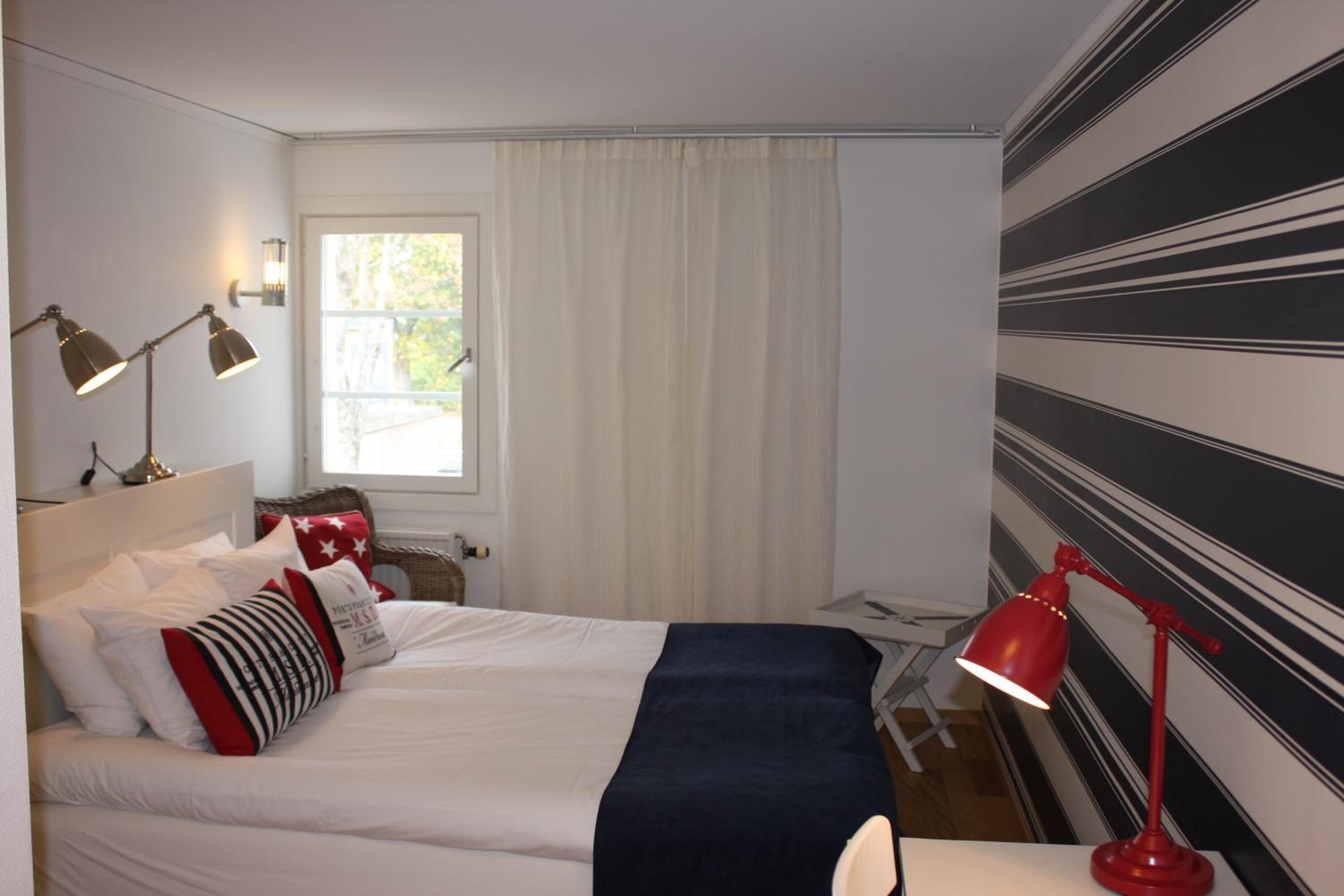Un dormitorio ordenado con cama, dos lámparas de noche, una ventana con cortinas y una pared a rayas.