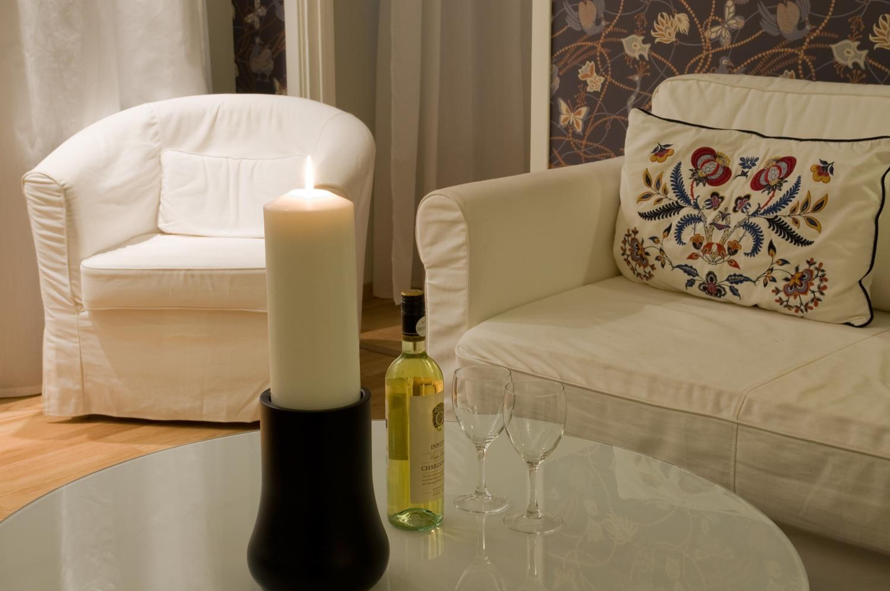 A white sofa with a floral pillow, a lit candle, a bottle of white wine, and two empty wine glasses on a round table.