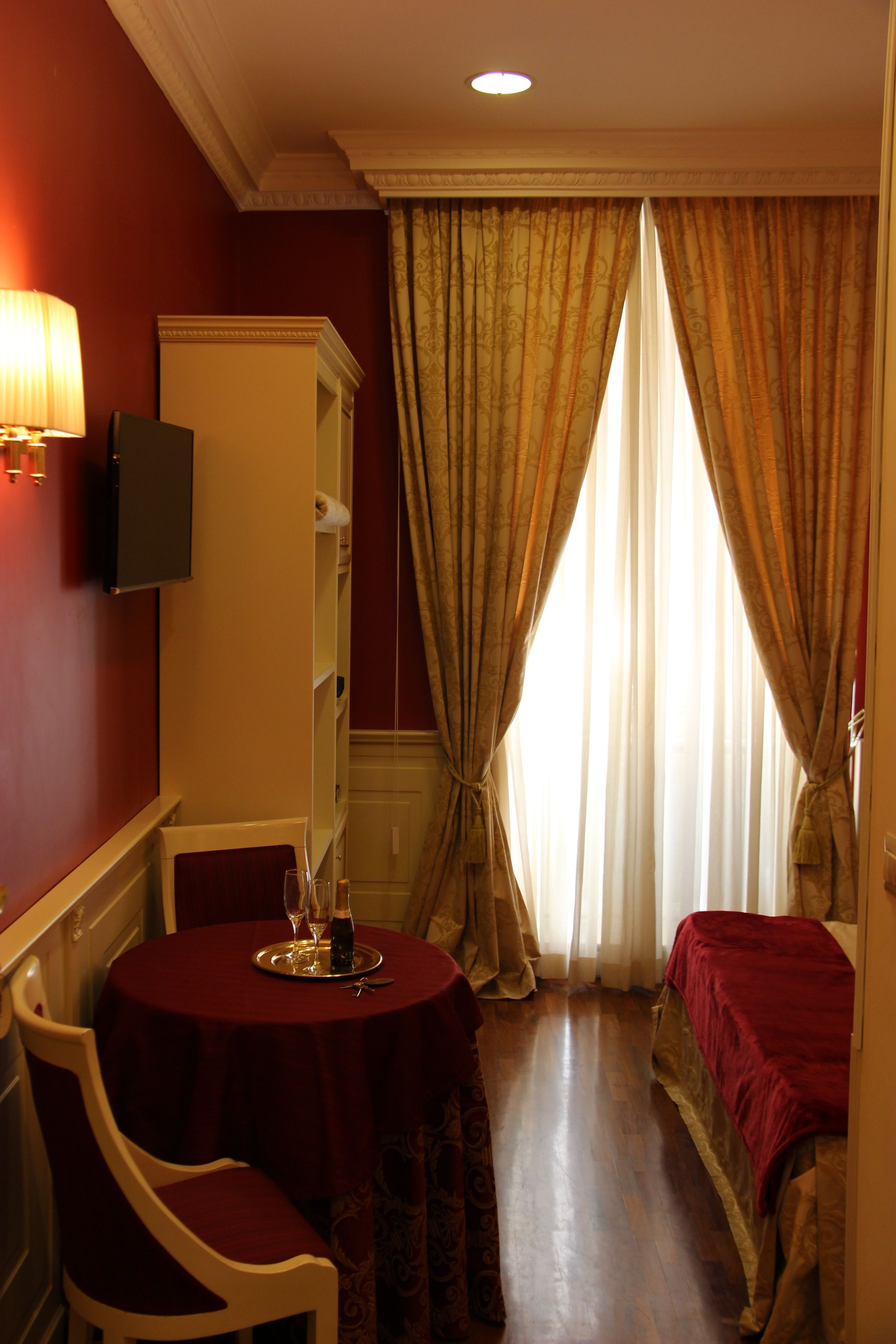 Ein Hotelzimmer mit rundem Tisch, einem Sessel, einem Bett mit roter Decke, einem Kleiderschrank und einem an der Wand montierten Fernseher.