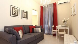 A modern living room with a grey sofa, red and grey cushions, a dining table with chairs, and a small white bench. There are framed pictures on the wall and a chandelier above the dining area. - Dimora Frattina (Rome)