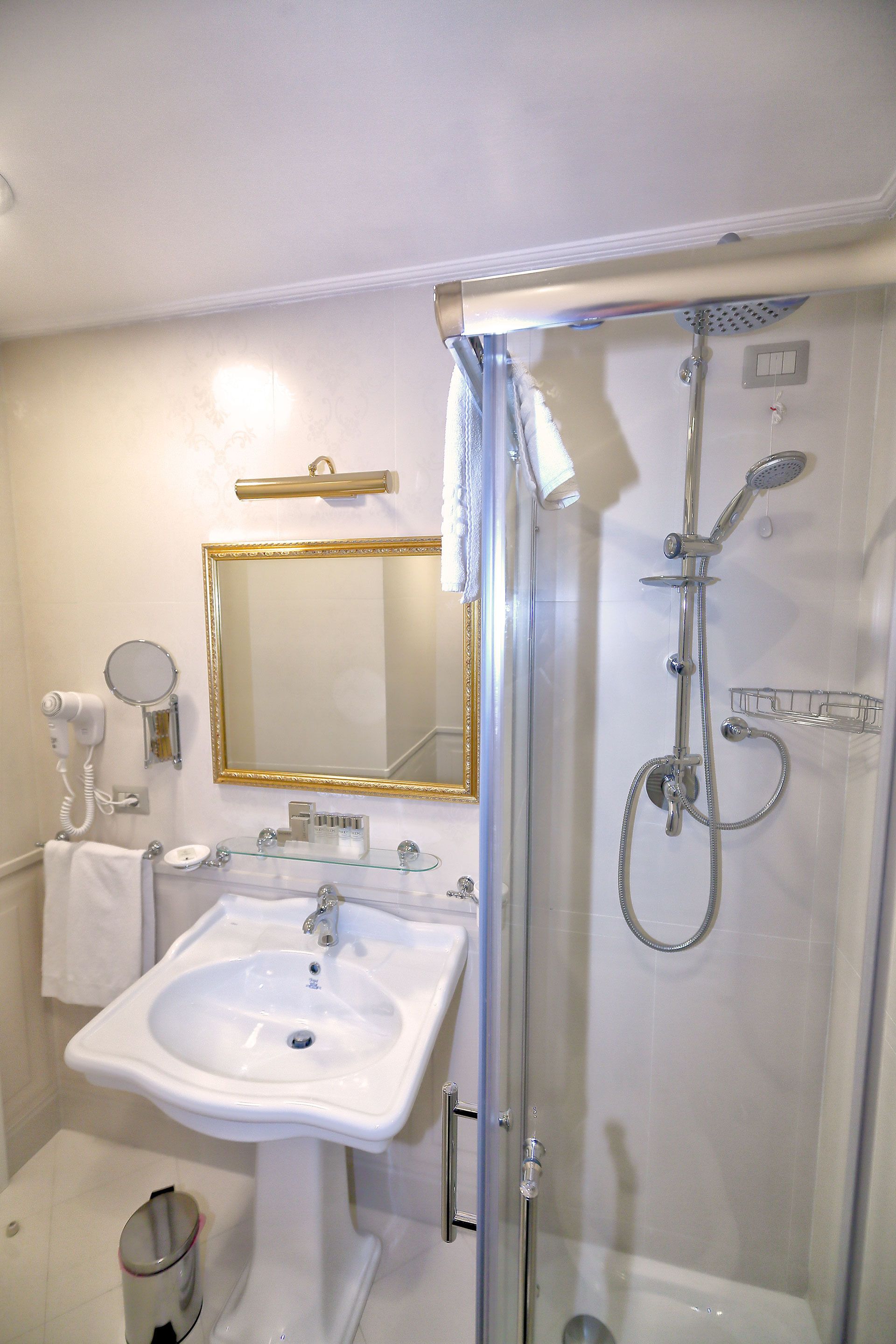 Casa de banho com uma pia branca, um espelho com moldura dourada, um secador de cabelo manual e uma cabine de duche com parede de vidro.