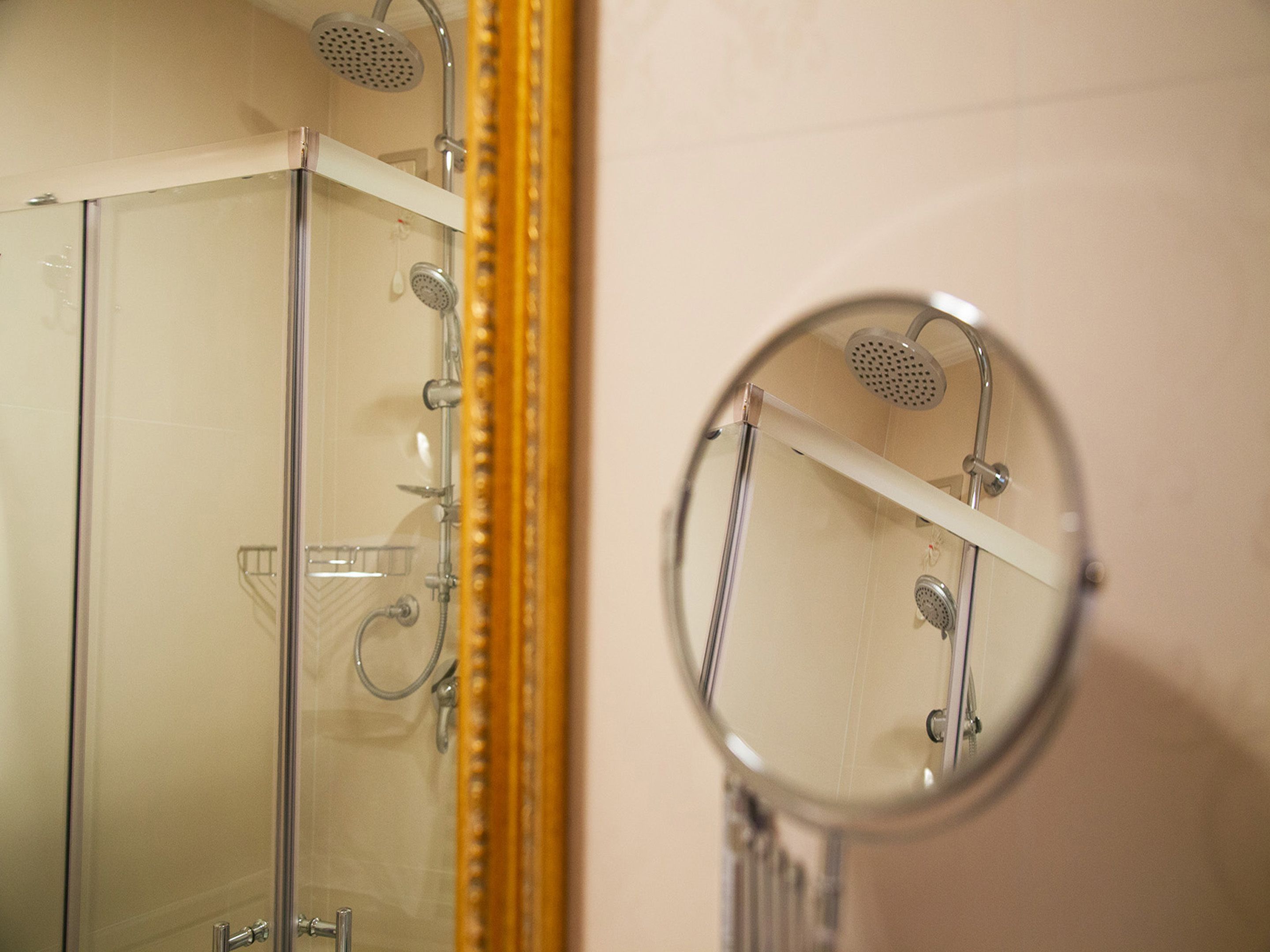 A bathroom with a glass shower enclosure, a white shelf above the shower, and a round mirror on the wall.