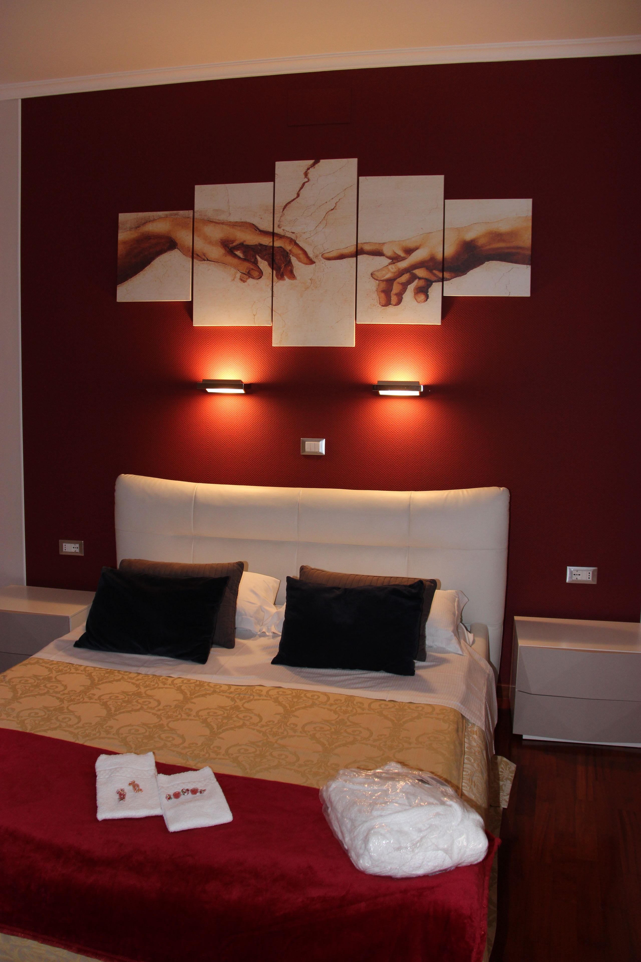 A hotel room with a large bed, two nightstands, a red wall, and a painting of hands reaching towards each other.