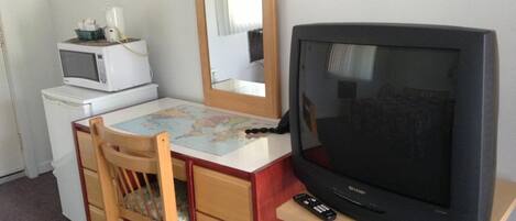 Una habitación compacta con escritorio de madera, televisión, microondas, refrigerador pequeño y espejo.