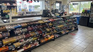 Interior de una tienda de comestibles con estantes llenos de productos, una caja registradora y un estacionamiento visible afuera.