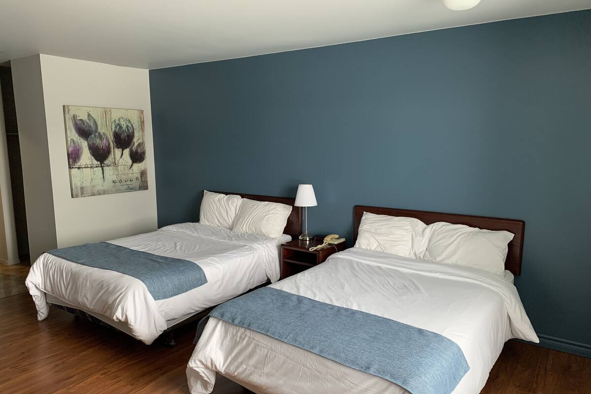 A hotel room with two beds, a nightstand with a lamp and phone, a painting on the wall, and wooden flooring.