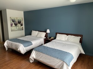 A hotel room with two beds, a nightstand with a lamp and phone, a painting on the wall, and wooden flooring.