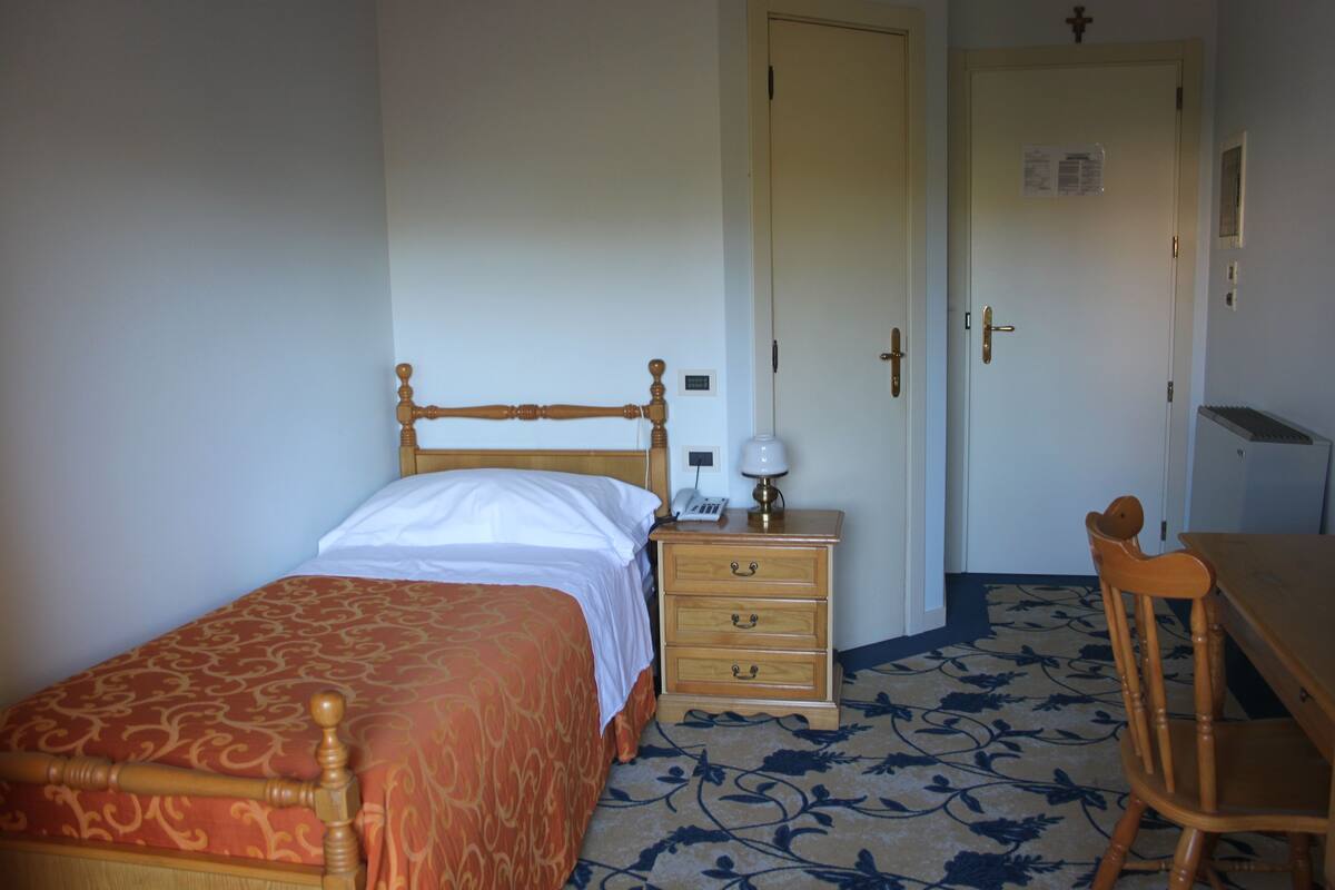 A bedroom with a bed, a nightstand with a lamp, a wooden chair, and a door with a sign.