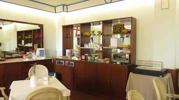A dining area with a buffet setup, including a counter with food items and a refrigerator. There are wicker chairs and a table with a white tablecloth.