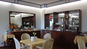 Un coin repas avec une table dressée pour quatre personnes, entourée de chaises en osier. On aperçoit, au fond, un espace cuisine avec divers objets rangés sur des étagères.