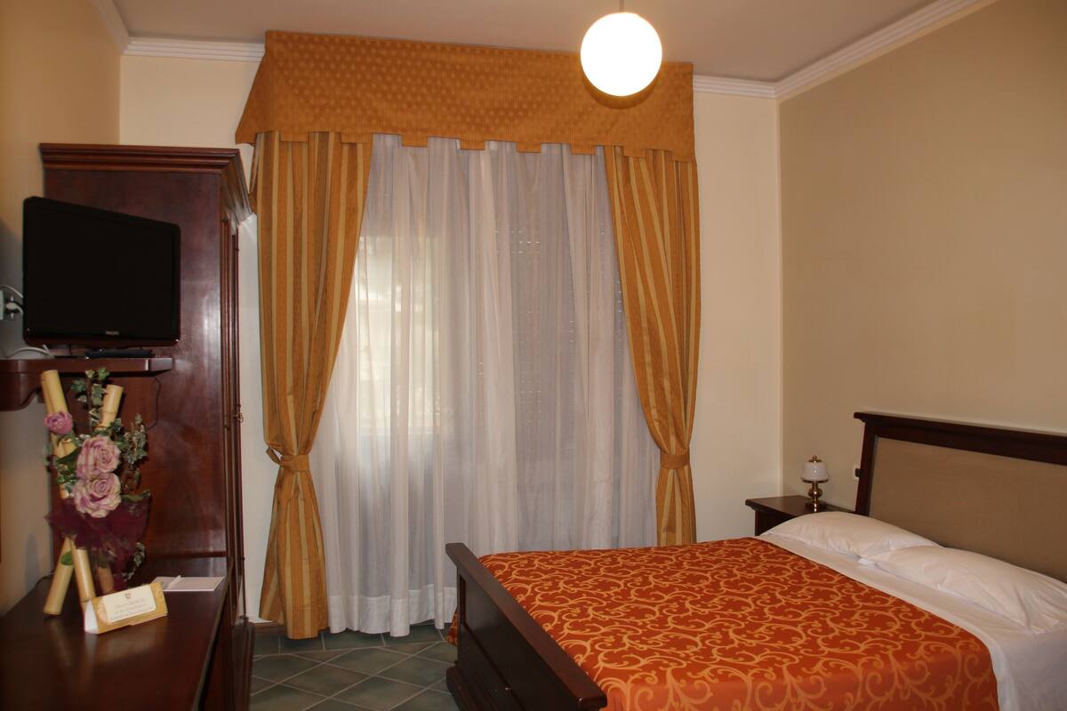 A hotel room with a bed, a TV on a wooden stand, a nightstand with a lamp, and a window with curtains.